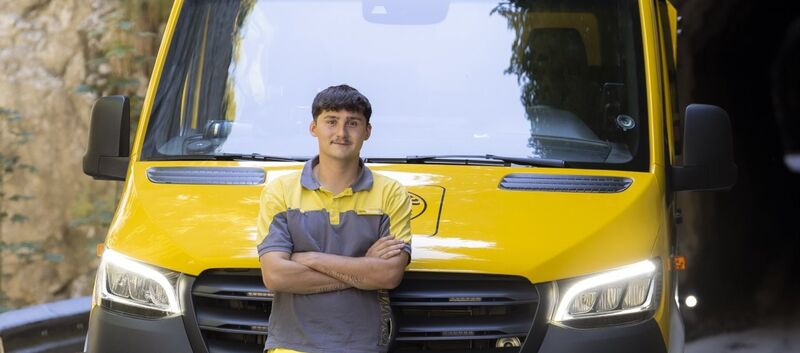Auf dem Bild sieht man einen jungen ÖAMTC Mitarbeiter. er lehnt mit verschränkten Armen an einen gelben Klein LKW.