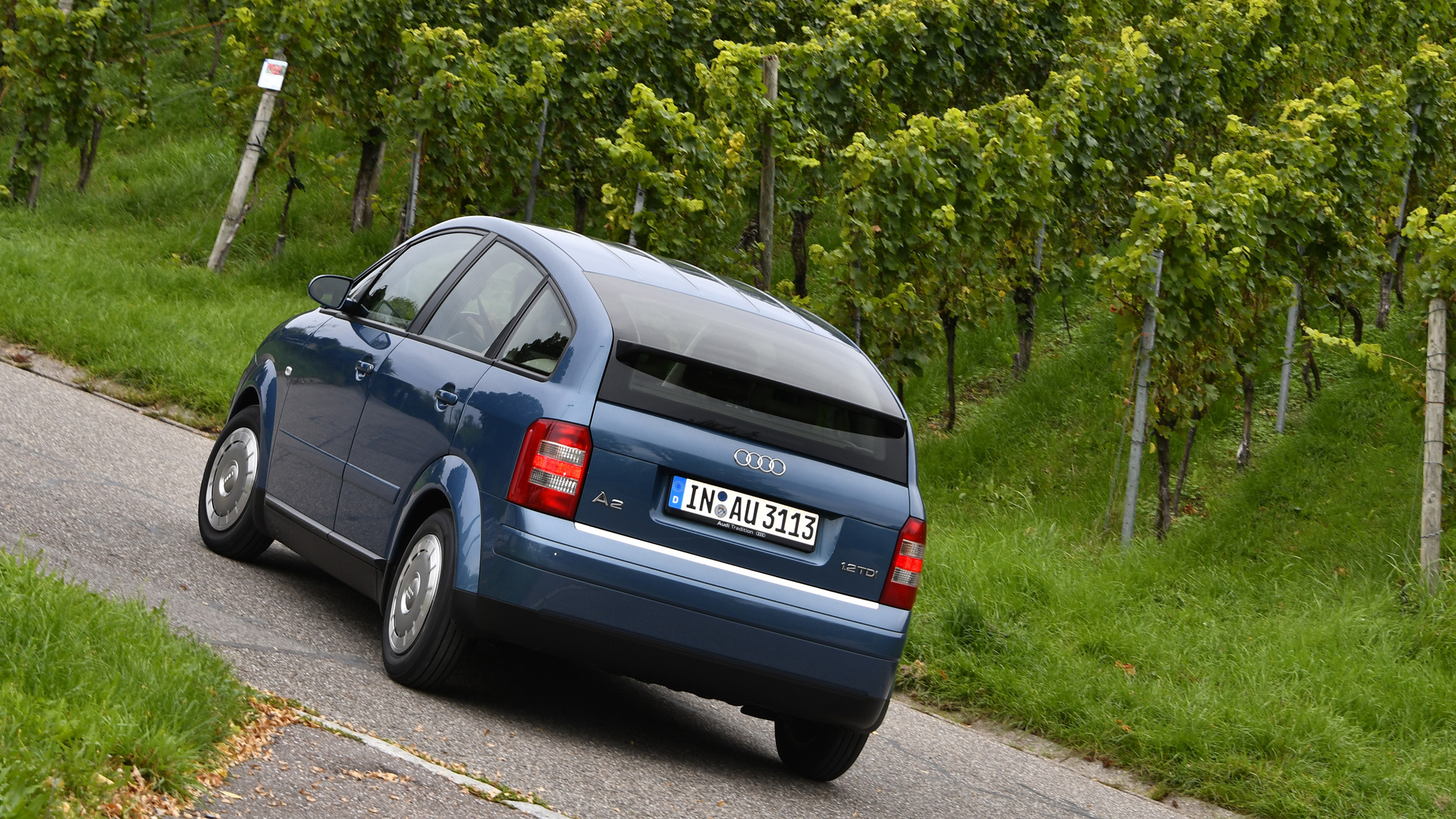 Heck eines alten, blauen Audi A2, der um eine Kurve fährt.