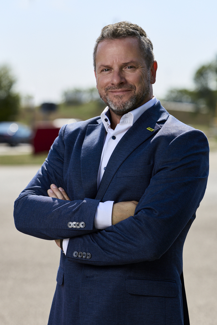 Porträt von Andreas Rouschal in einem blauen Anzug mit verschränkten Händen in die Kamera lächelnd, im Hintergrund sieht man ein Fahrtechnik-Training