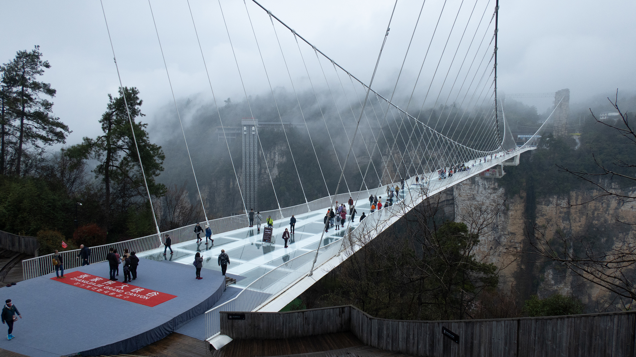 Menschen gehen über eine gläserne Fußgänger-Hängebrücke in einer nebligen Schlucht.