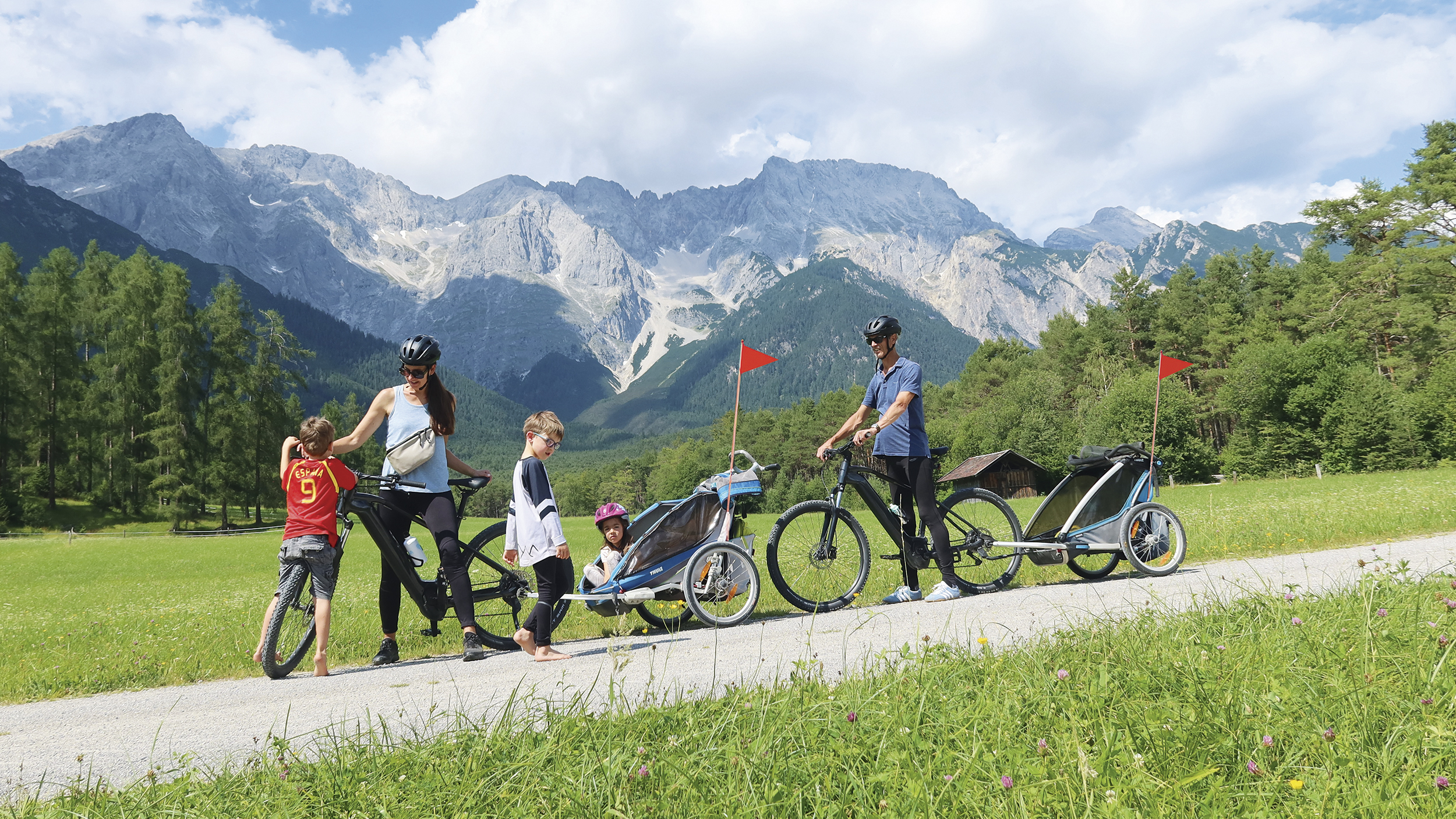 Eine Familie mit 3 Kleinkindern ist mit 2 Radanhängern unterwegs. Im Hintergrund die Bergkulisse.