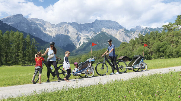 Eine Familie mit 3 Kleinkindern ist mit 2 Radanhängern unterwegs. Im Hintergrund die Bergkulisse.