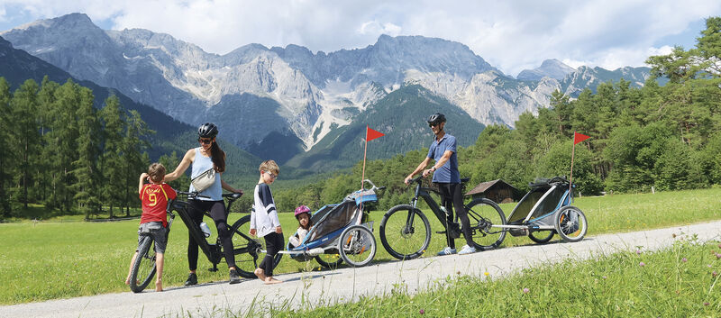 Eine Familie mit 3 Kleinkindern ist mit 2 Radanhängern unterwegs. Im Hintergrund die Bergkulisse.