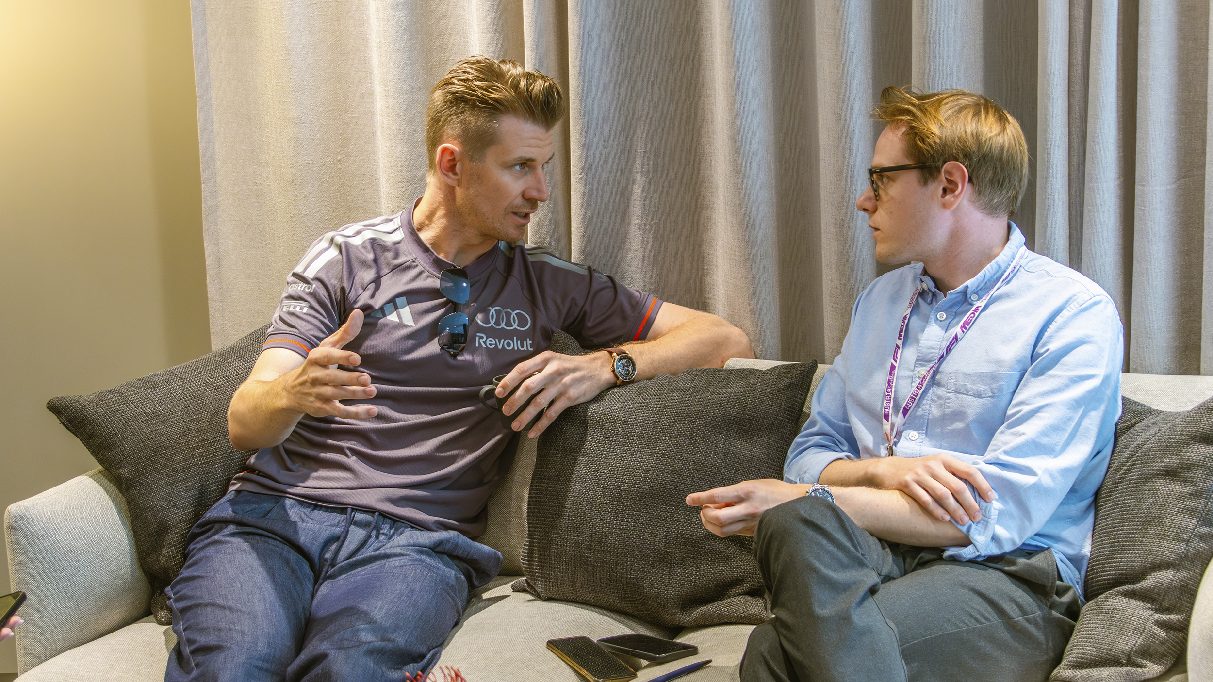 Nico Hülkenberg spricht auf einem Sofa sitzend mit einem Mann in hellblauem Hemd und Brille.