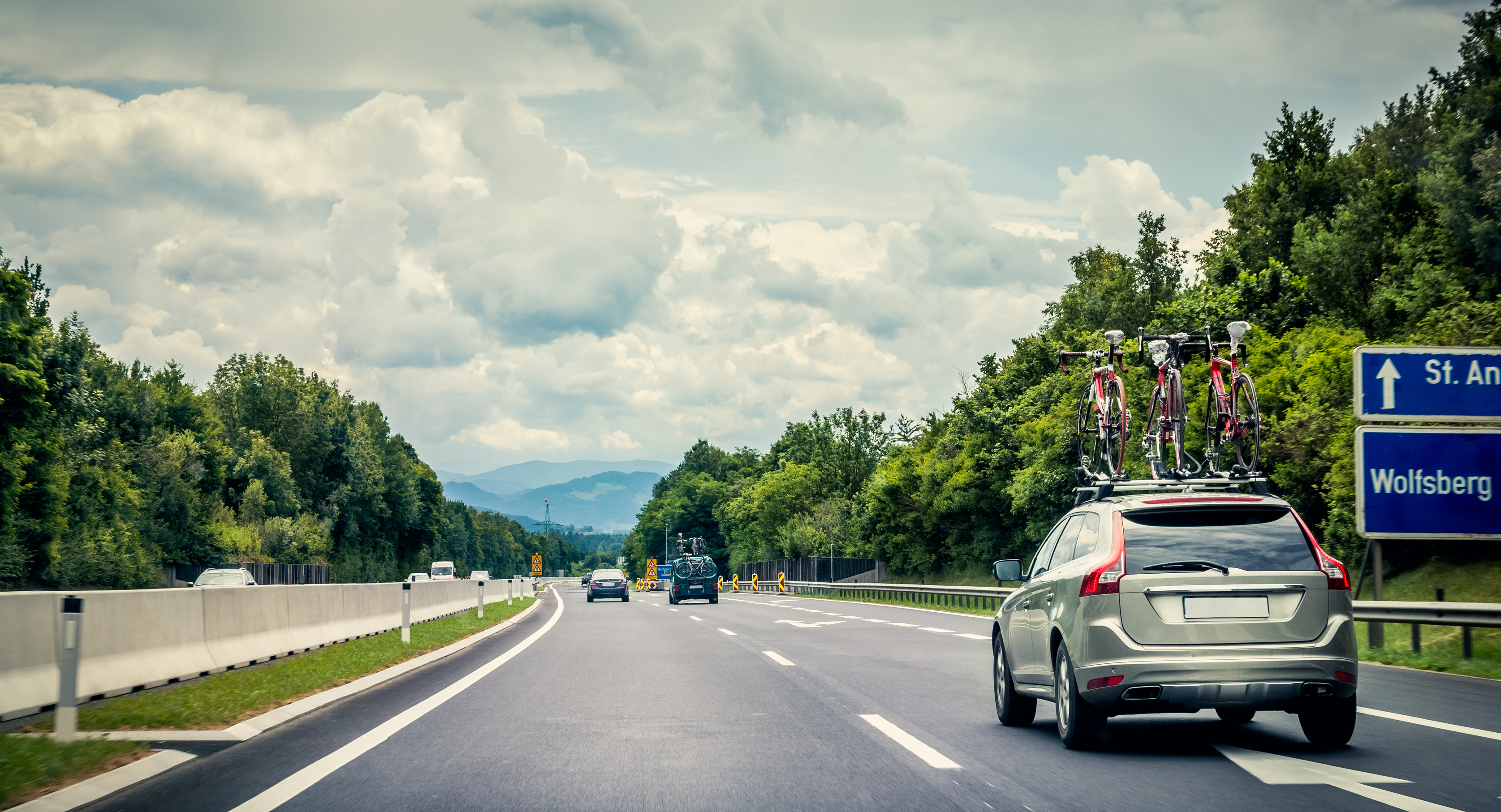 Auf einer Autobahn fährt ein graues Auto mit drei Fahrrädern am Dachträger. Neben der Straße sind grüne Bäume und der Himmel ist bewölkt.