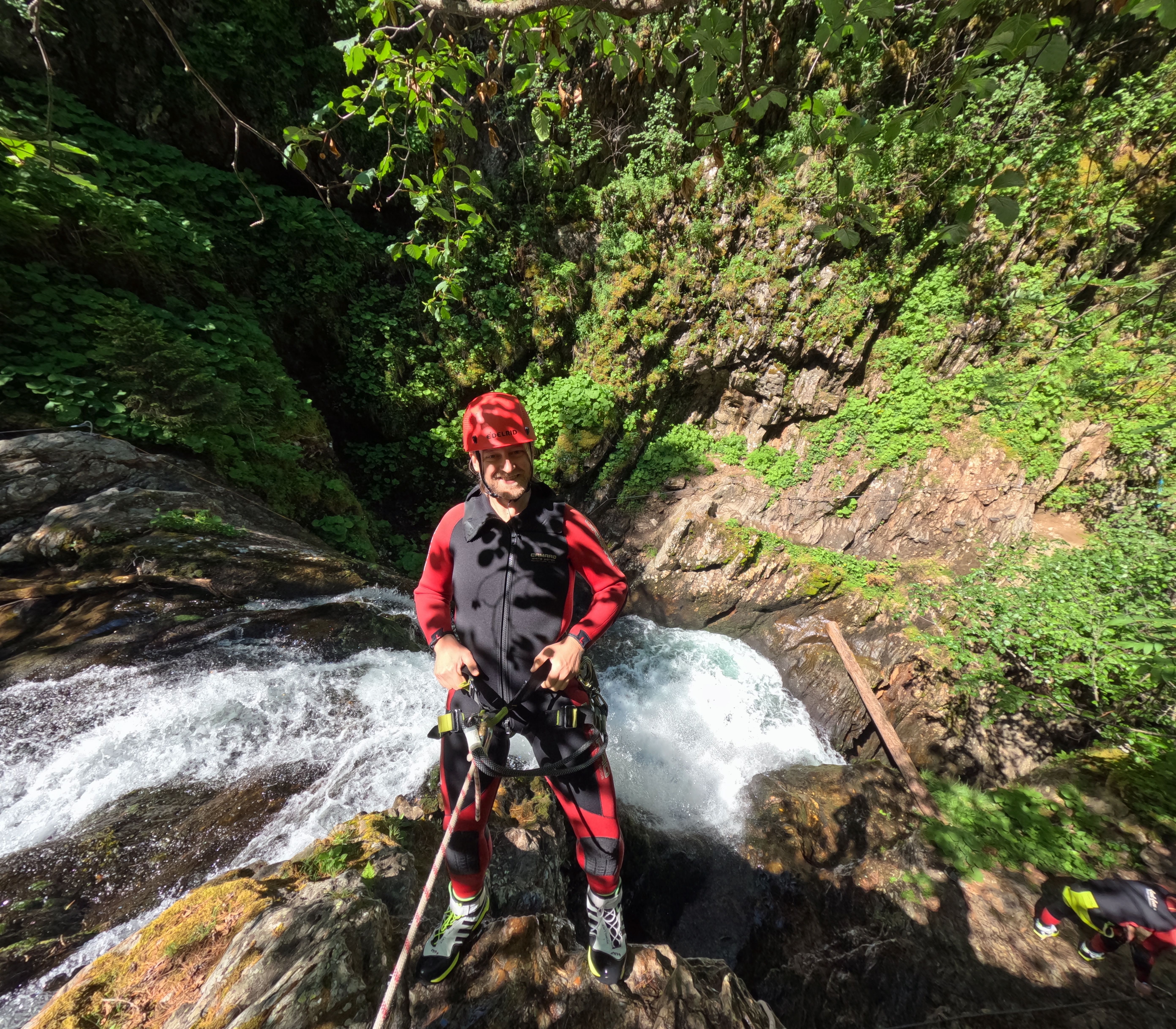 Es ist ein Mann in Neoprenanzu zu sehen, der sich gerade an einem Wasserfall abseilt. Er hat Spaß und lacht. 