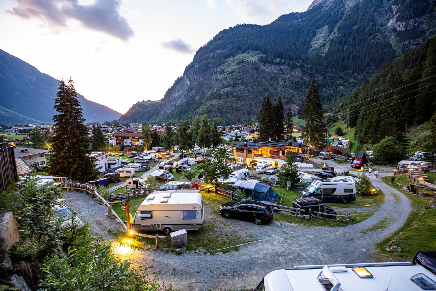 Man sieht auf den Bild einen Campingplatz bei leichter Dämmerung. Im Hitnergrund sieht man Berge und viel Natur. 