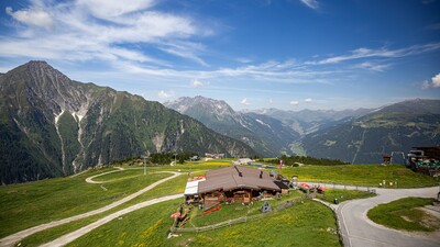 Das Bild zeigt eine grüne Wiese, eine Almhütte und eine Bergkette.