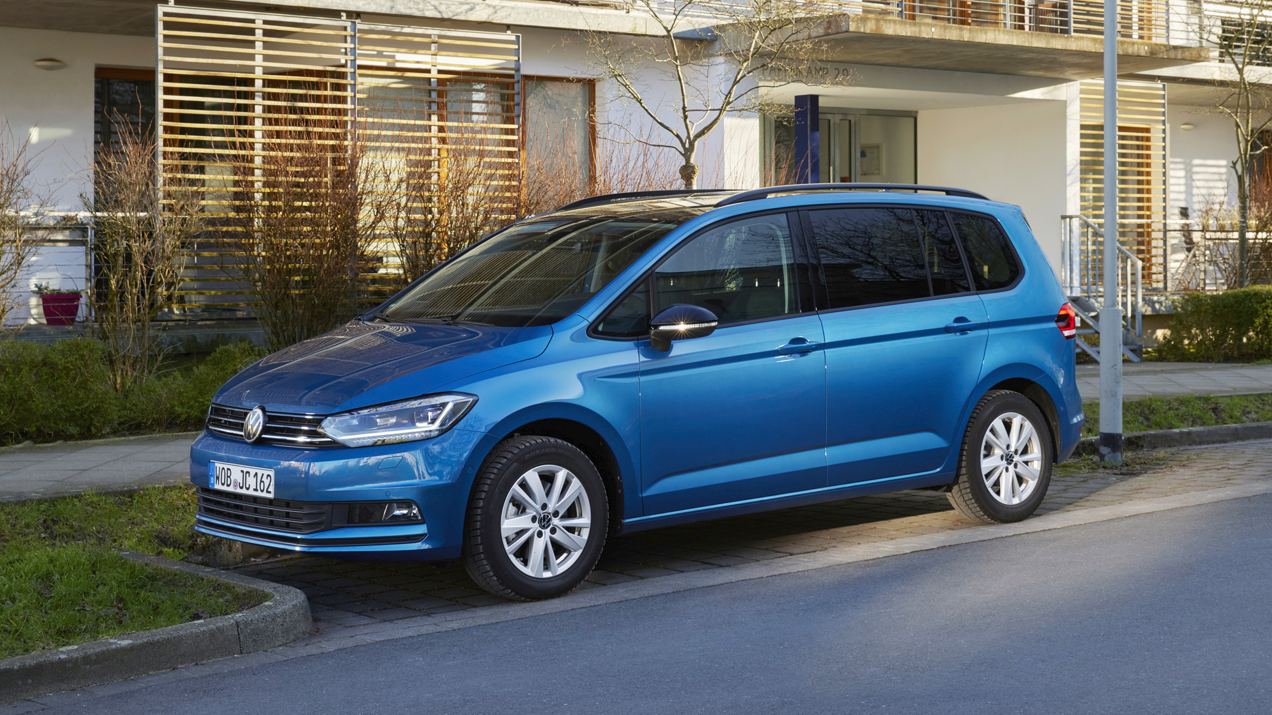 Blauer VW Touran, Kompaktvan mit Dachreling in Halbfrontansicht von links schräg vorn, am Straßenrand vor einem modernen Wohnhaus mit Holzlamellen geparkt.