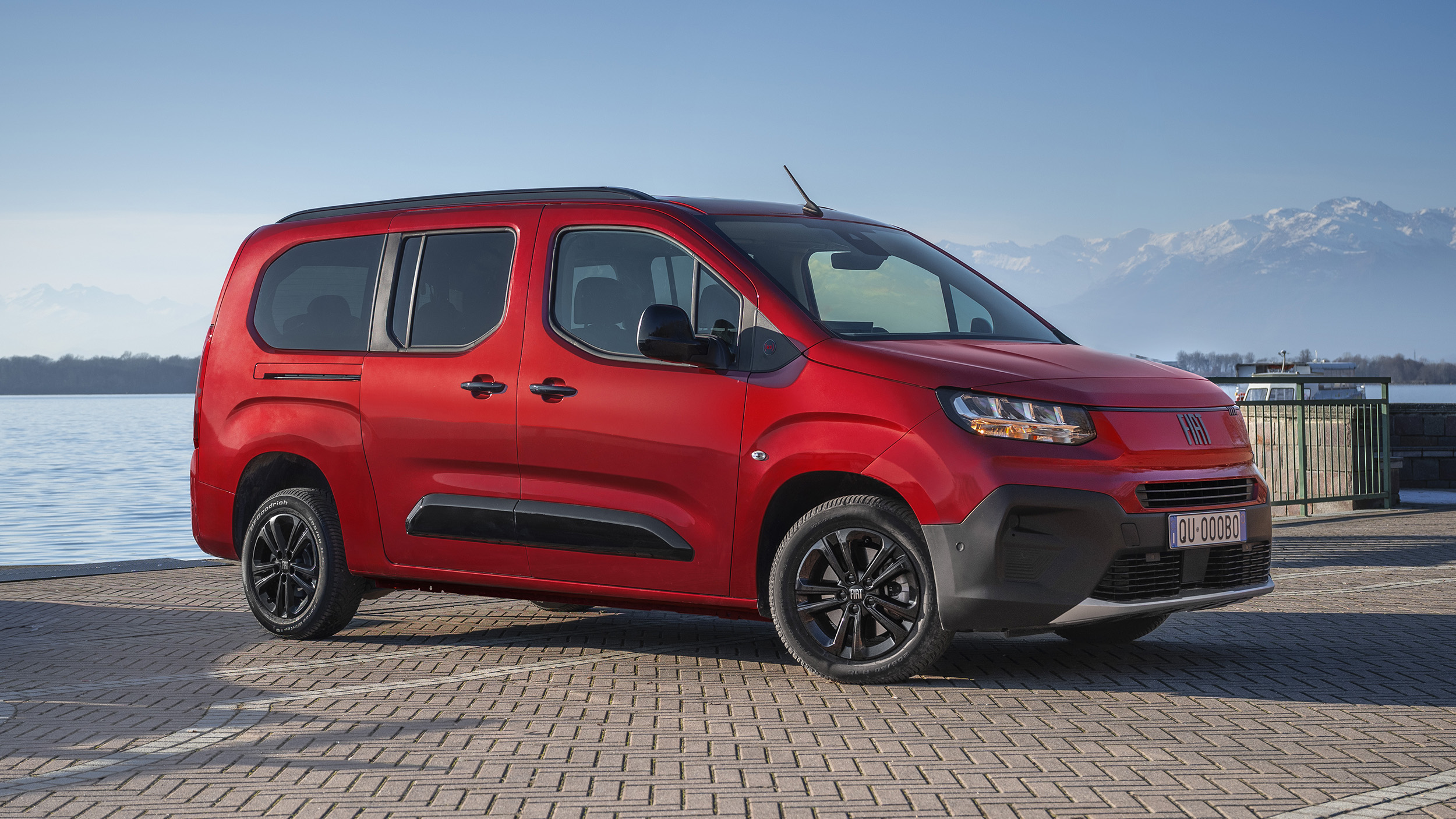 Roter Fiat Qubo L, Hochdachkombi mit Schiebetür und schwarzen Seitenleisten, in Halbfrontansicht von rechts schräg vorn auf einer Uferpromenade; im Hintergrund See und schneebedeckte Berge.