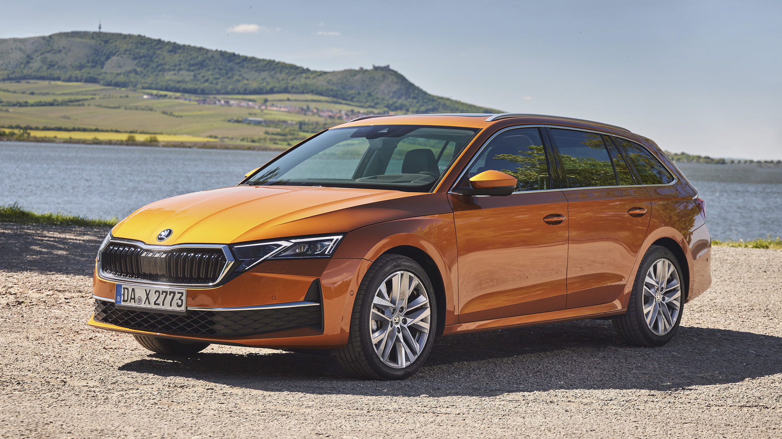 Orangefarbener Škoda Octavia Combi, Kombi in Halbfrontansicht von links schräg vorn, auf einem Schotterplatz an einem See; dahinter Weinberge und ein Hügel.
