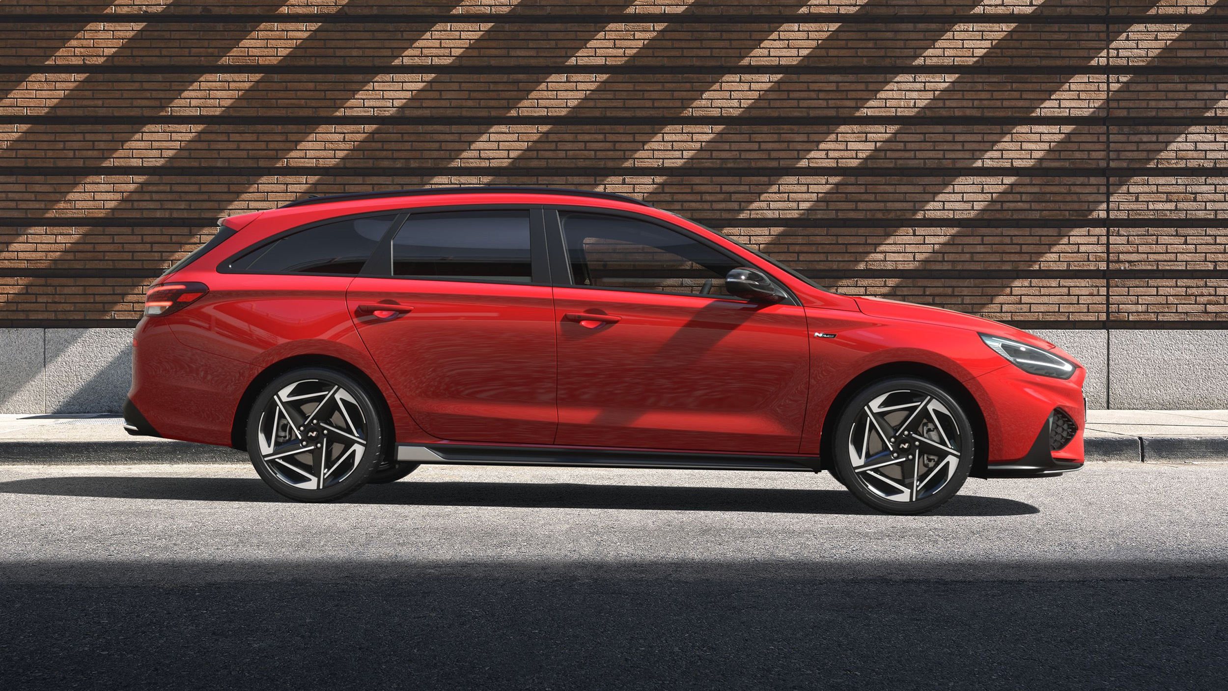 Roter Hyundai i30 Kombi in vollständiger Seitenansicht von rechts, mit Dachreling und mehrspeichigen Leichtmetallfelgen, an einer Ziegelwand mit Schattenmuster geparkt.
