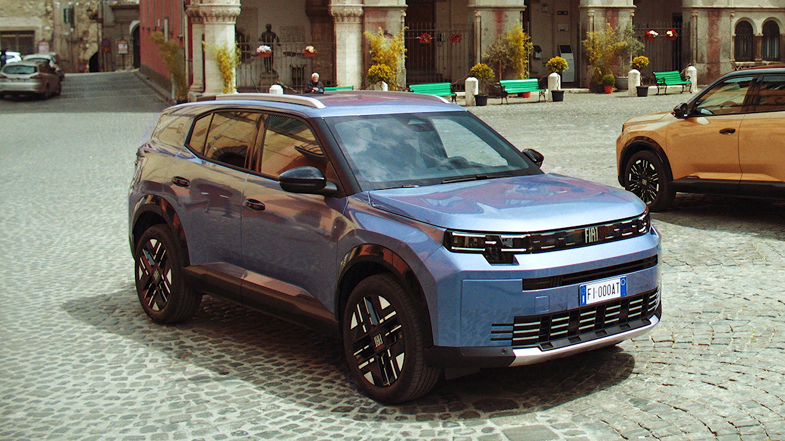 Dunkelblauer DacBlaugrauer Fiat Grizzly, SUV in Halbfrontansicht von rechts schräg vorn, auf einem gepflasterten Platz einer italienischen Altstadt; rechts im Bild ein orangefarbenes Schwestermodell, im Hintergrund historische Arkaden.a Bigster Journey, SUV mit Dachreling und Kunststoffbeplankung, in Halbfrontansicht von links schräg vorn vor einer Betonwand; im Hintergrund bewaldeter Hügel.
