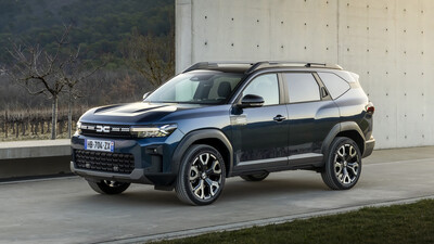 Dunkelblauer Dacia Bigster Journey, SUV mit Dachreling und Kunststoffbeplankung, in Halbfrontansicht von links schräg vorn vor einer Betonwand; im Hintergrund bewaldeter Hügel.