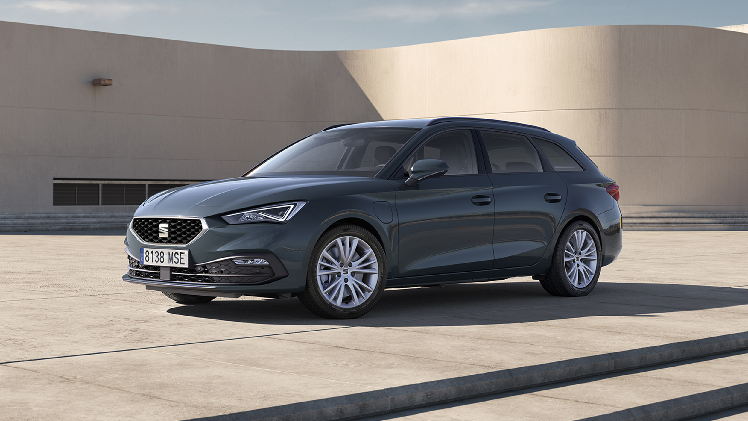 Dunkelgrauer Seat Leon Sportstourer, Kombi in Halbfrontansicht von links schräg vorn, auf einer Betonfläche vor einer hellen, geschwungenen Gebäudewand.