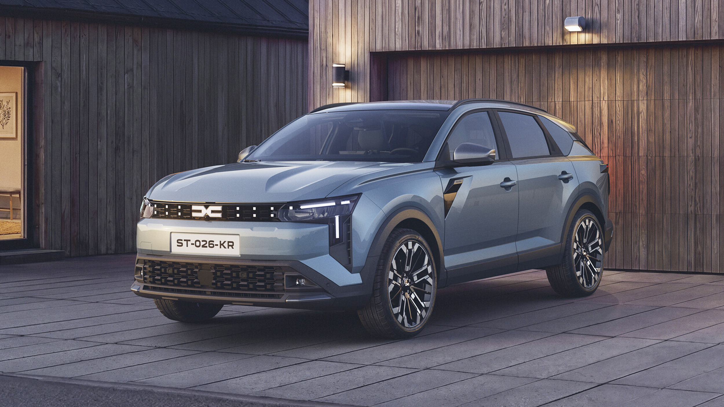 Hellblaugrauer Dacia Striker Hybrid 155, SUV in Halbfrontansicht von links schräg vorn, vor einer holzverschalten Hausfassade in der Dämmerung; Tagfahrlicht und Leuchtsignatur sind eingeschaltet.