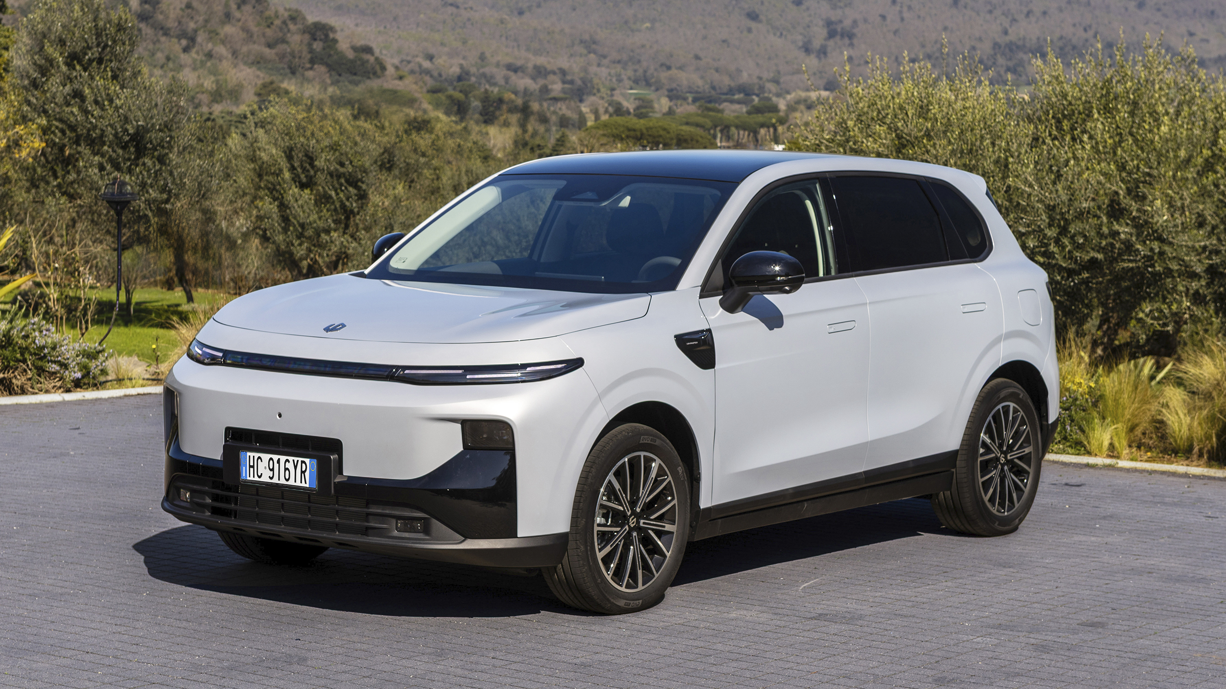 Weißer Leapmotor B10 mit schwarzem Dach, SUV in Halbfrontansicht von links schräg vorn, auf gepflastertem Vorplatz zwischen Olivenbäumen; im Hintergrund bewaldete Hügel.