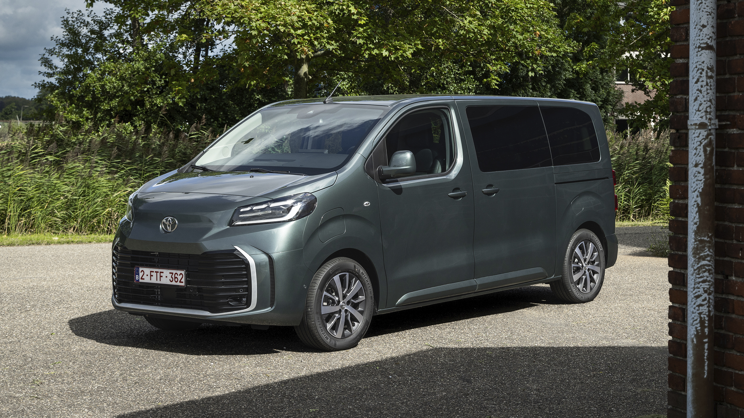 Graugrüner Toyota Proace Verso, Van mit Schiebetür und verglaster zweiter Sitzreihe, in Halbfrontansicht von links schräg vorn auf einem Asphaltplatz; im Hintergrund Bäume und Schilf.