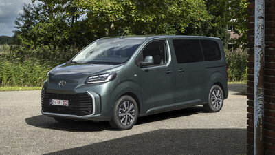 Graugrüner Toyota Proace Verso, Van mit Schiebetür und verglaster zweiter Sitzreihe, in Halbfrontansicht von links schräg vorn auf einem Asphaltplatz; im Hintergrund Bäume und Schilf.