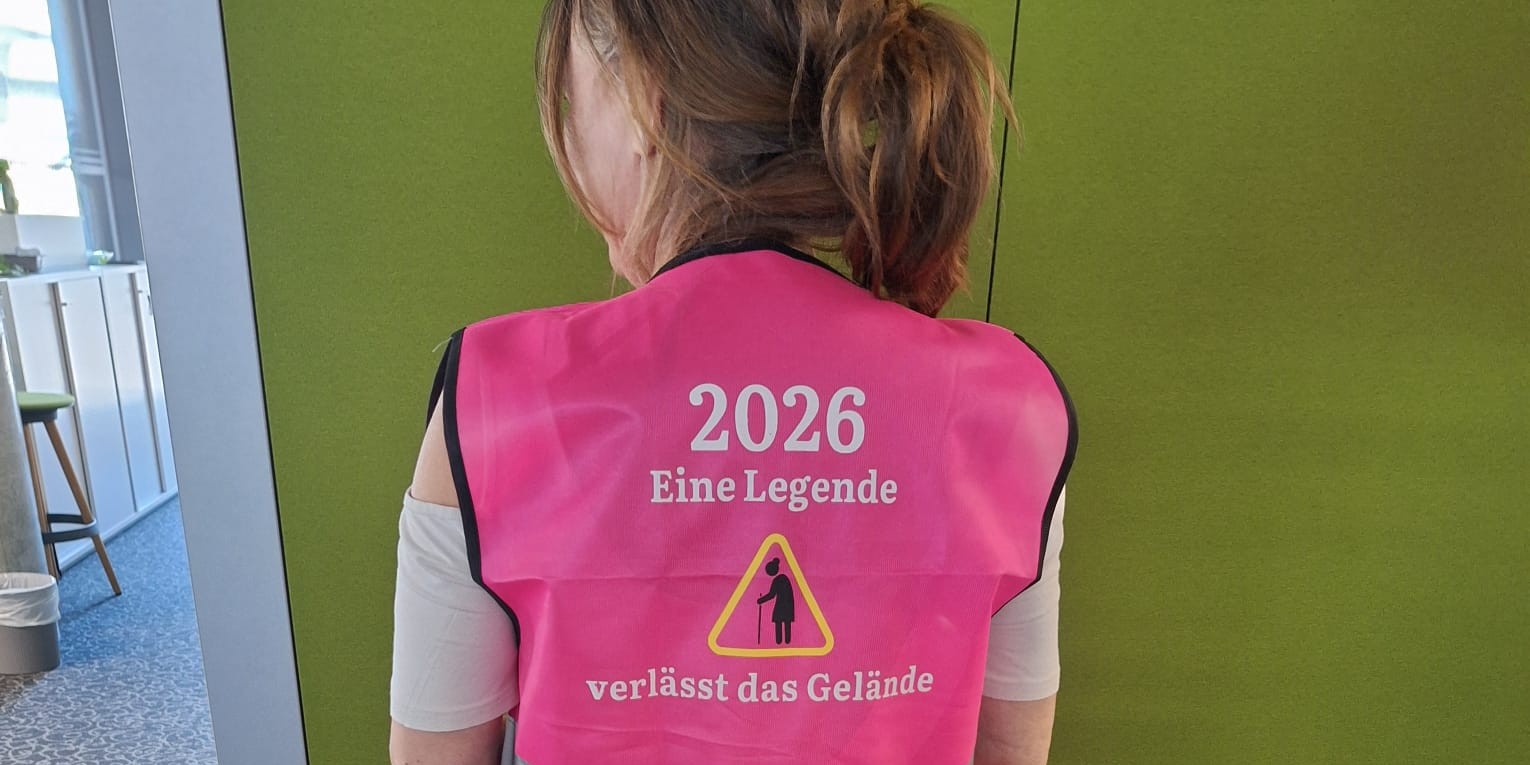 Eine Person steht vor einer grünen Wand. Sie trägt eine rosa warnweste. Auf dieser steht. 2026. Eine Legende verlässt das Gelände