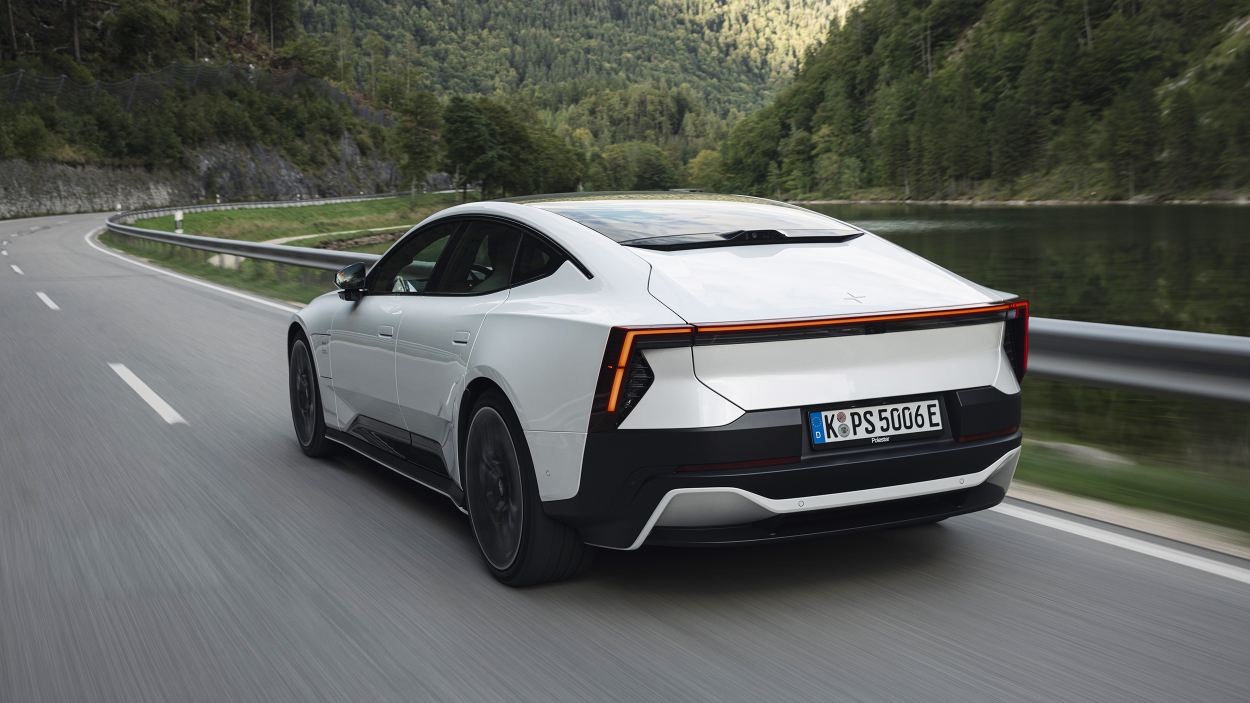 Weißer Polestar 5 Performance fährt auf einer kurvigen Straße neben einem See und bewaldeten Bergen.