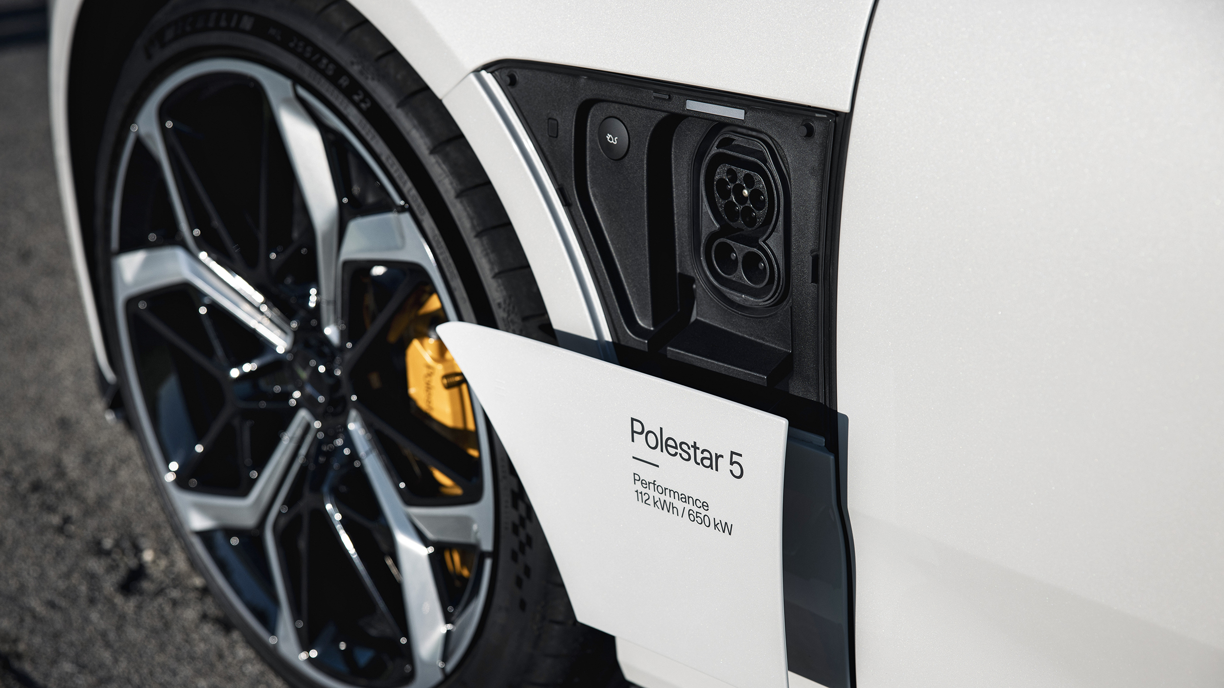 Geöffnete Ladeklappe eines weißen Polestar 5 mit CCS-Ladeanschluss neben einem schwarzen Leichtmetallrad und gelbem Bremssattel.