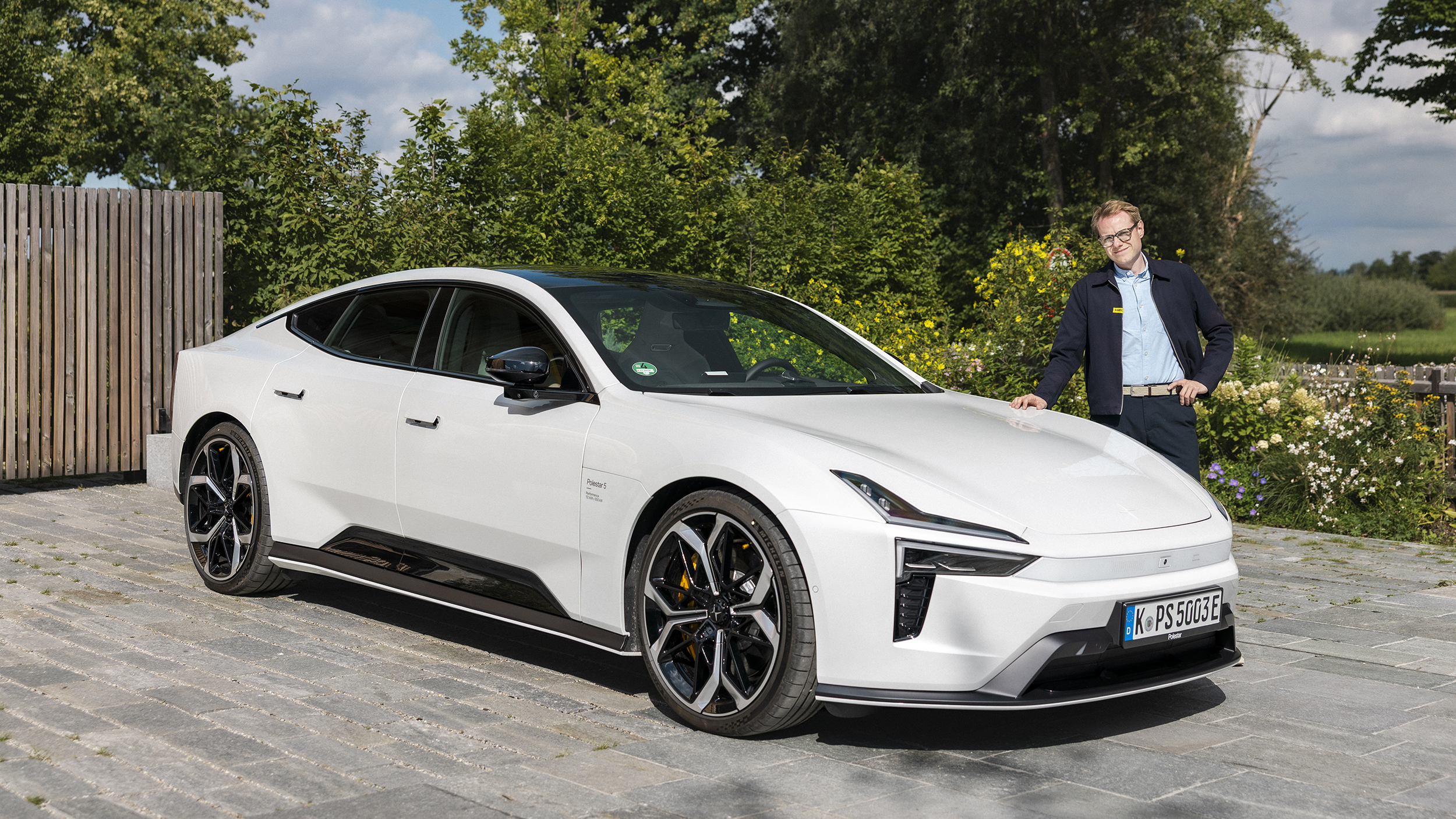 Weißer Polestar 5 steht auf einer gepflasterten Einfahrt vor einem Haus; daneben steht ein Mann mit Brille.