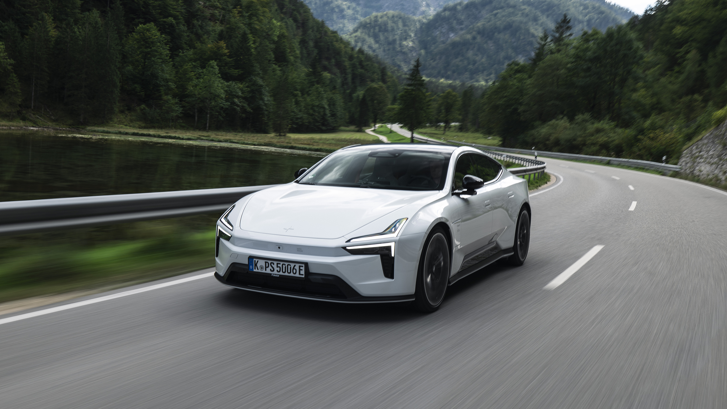 Weißer Polestar 5 Performance fährt auf einer kurvigen Straße neben einem See und bewaldeten Bergen.