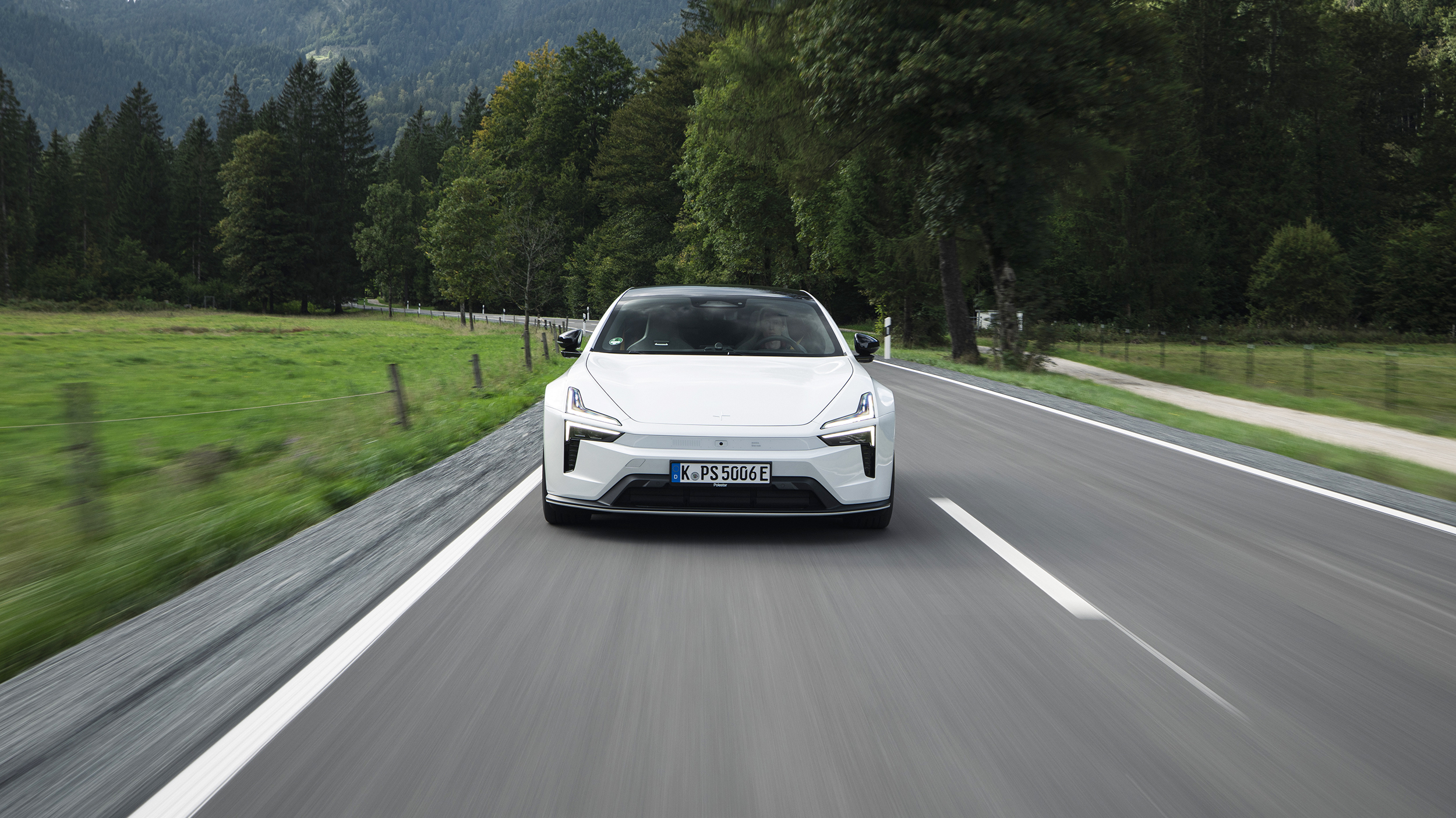 Weißer Polestar 5 fährt frontal auf einer Landstraße durch eine grüne, bewaldete Landschaft.