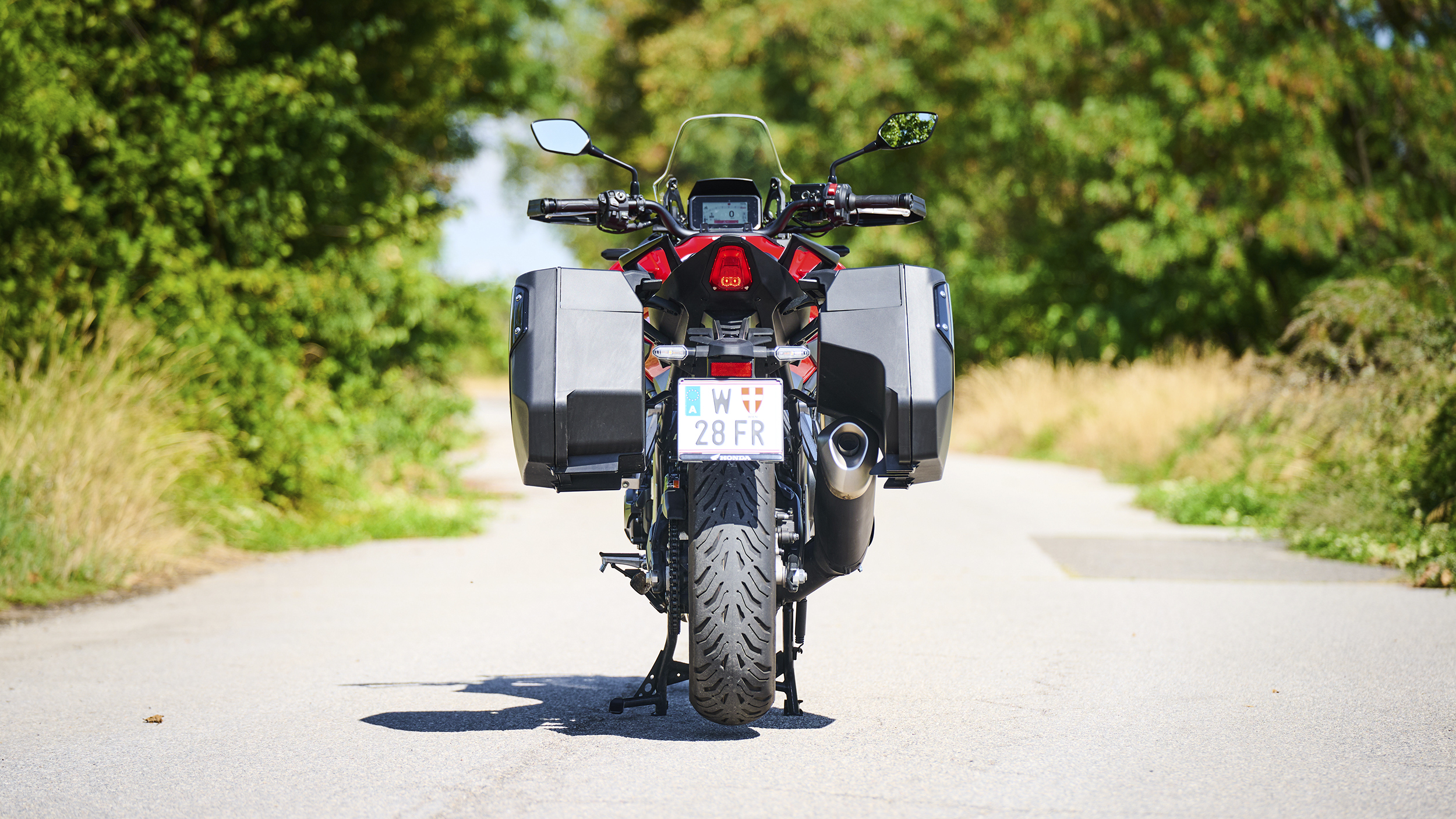 Rote Honda CB1000GT mit schwarzen Seitenkoffern, von hinten auf einer sonnigen Landstraße mit Bäumen am Straßenrand.