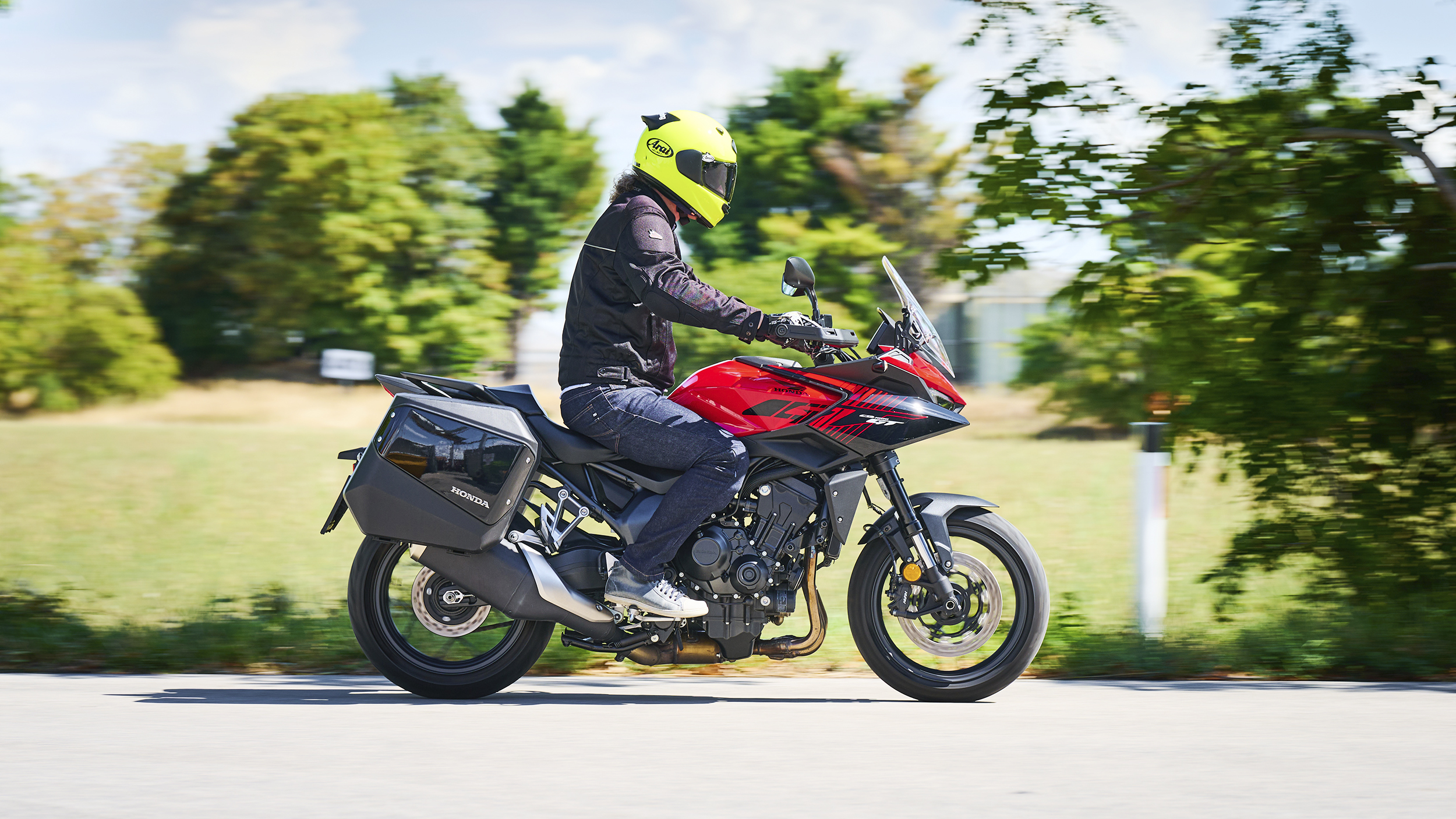 Motorradfahrerin oder Motorradfahrer mit gelbem Helm fährt eine rote Honda CB1000GT mit schwarzen Seitenkoffern auf einer Landstraße; der Hintergrund ist durch die Bewegung unscharf.