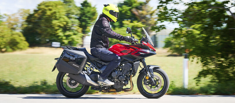 Motorradfahrerin oder Motorradfahrer mit gelbem Helm fährt eine rote Honda CB1000GT mit schwarzen Seitenkoffern auf einer Landstraße; der Hintergrund ist durch die Bewegung unscharf.