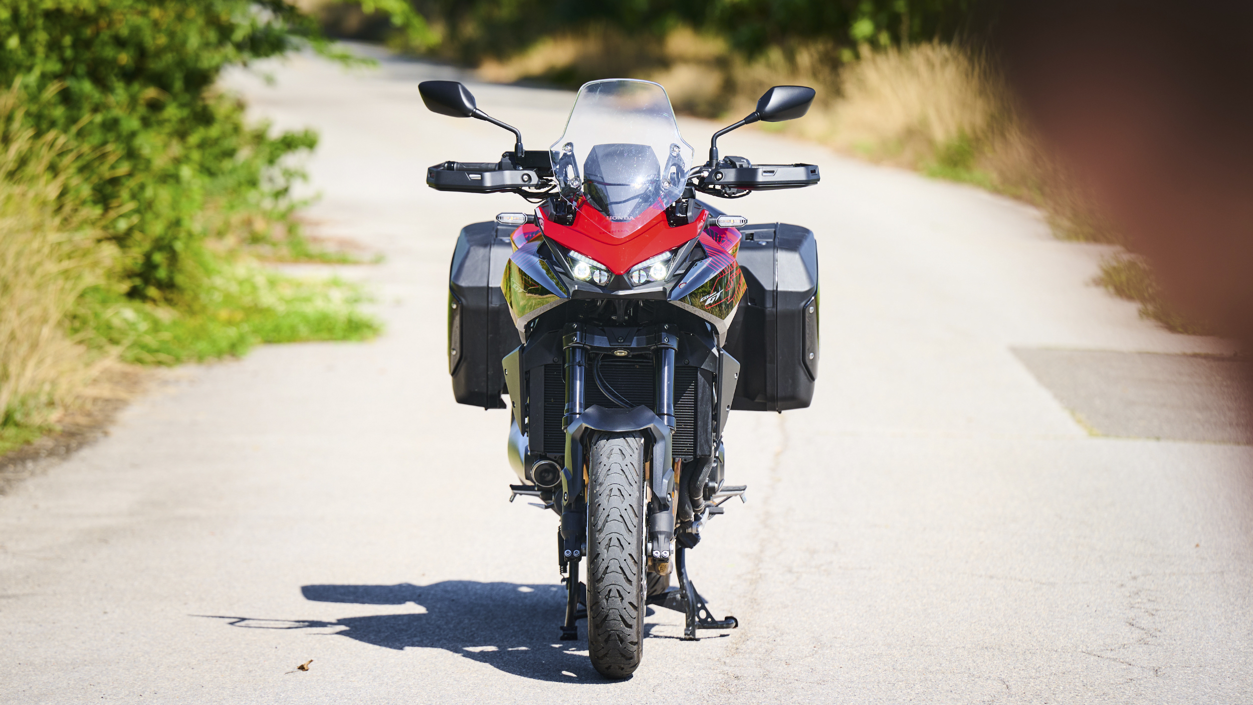 Rote Honda CB1000GT mit eingeschaltetem Doppelscheinwerfer und schwarzen Seitenkoffern in Frontansicht auf einer Landstraße.
