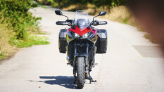 Rote Honda CB1000GT mit eingeschaltetem Doppelscheinwerfer und schwarzen Seitenkoffern in Frontansicht auf einer Landstraße.