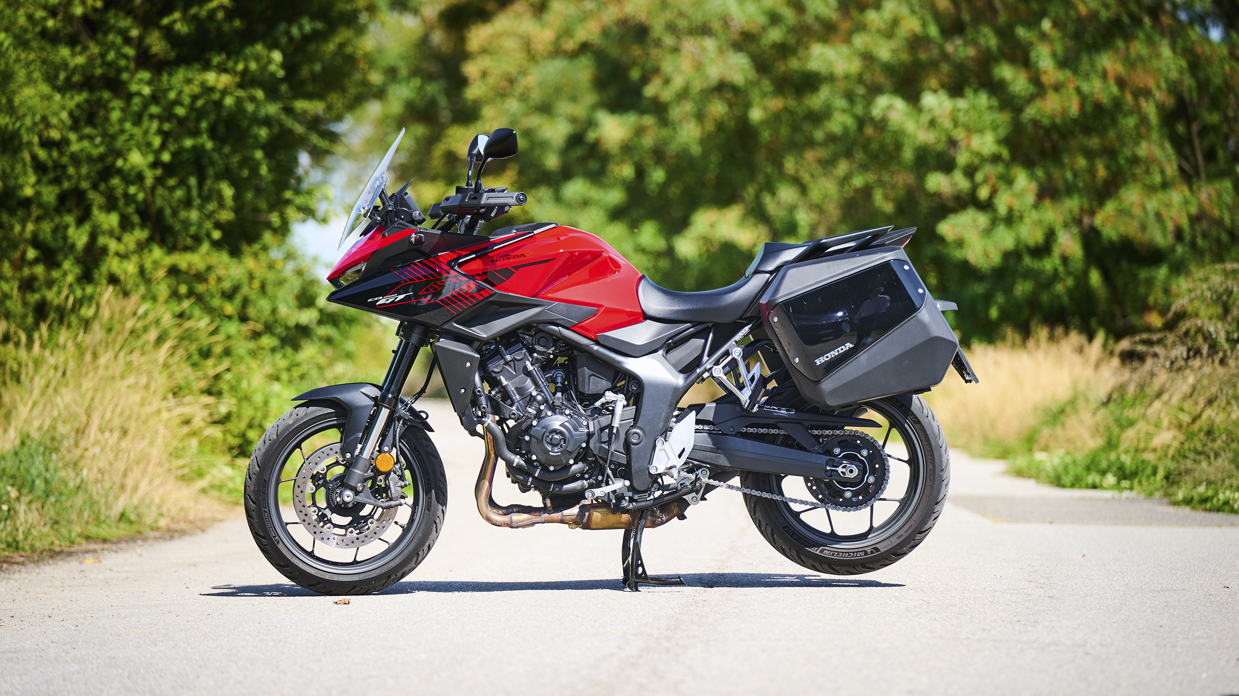 Rote Honda CB1000GT mit schwarzen Seitenkoffern in linker Seitenansicht, abgestellt auf einer sonnigen Landstraße.