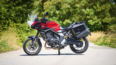 Rote Honda CB1000GT mit schwarzen Seitenkoffern in linker Seitenansicht, abgestellt auf einer sonnigen Landstraße.