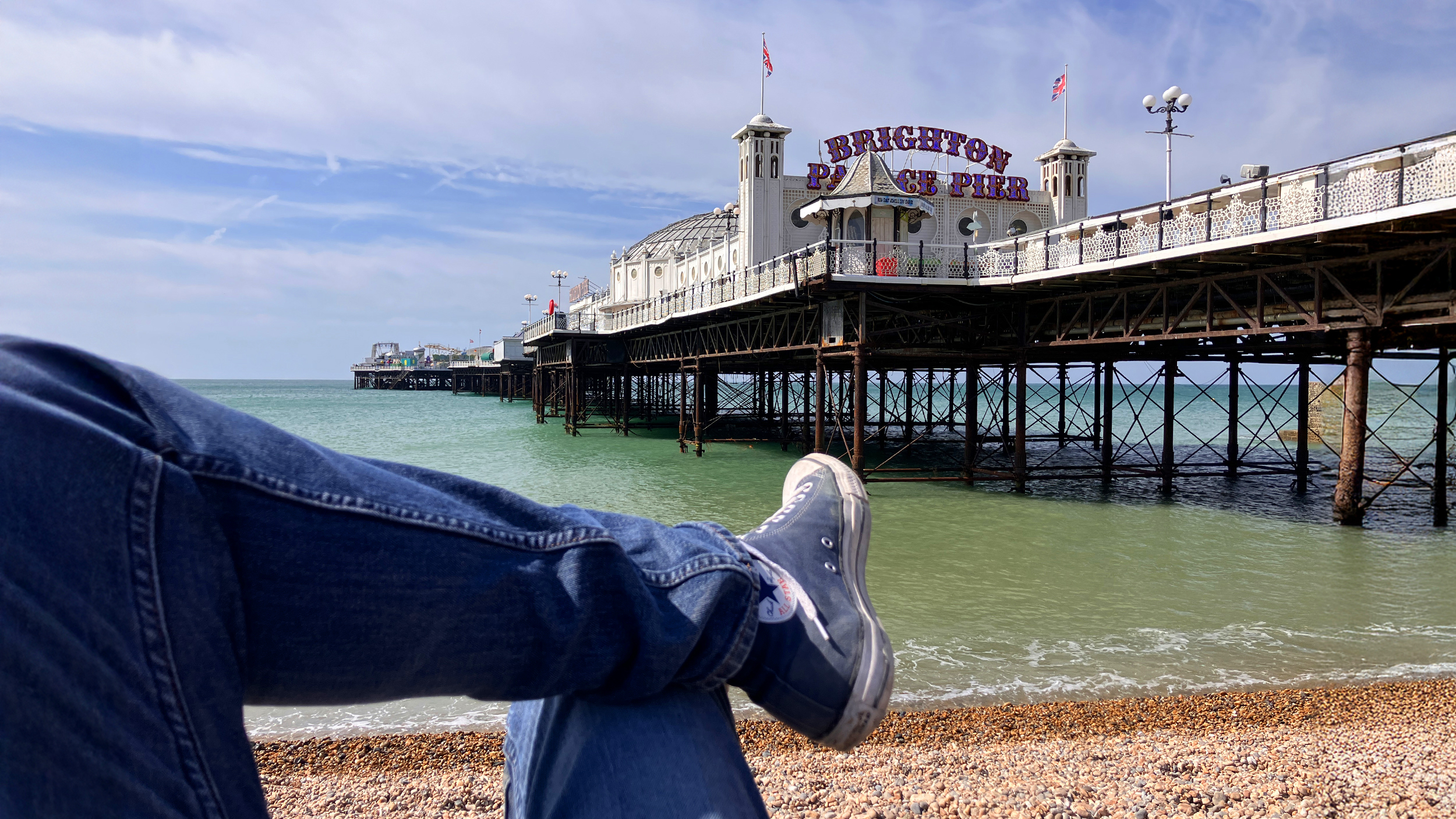 Blick vom Kiesstrand auf den Brighton Palace Pier; im Vordergrund liegen überkreuzte Beine in Jeans und Turnschuhen.