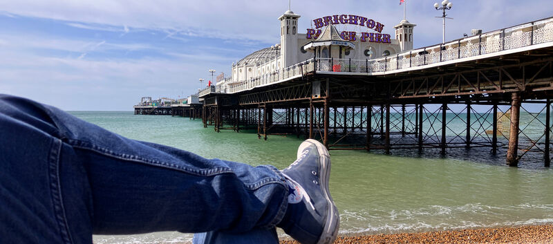 Blick vom Kiesstrand auf den Brighton Palace Pier; im Vordergrund liegen überkreuzte Beine in Jeans und Turnschuhen.