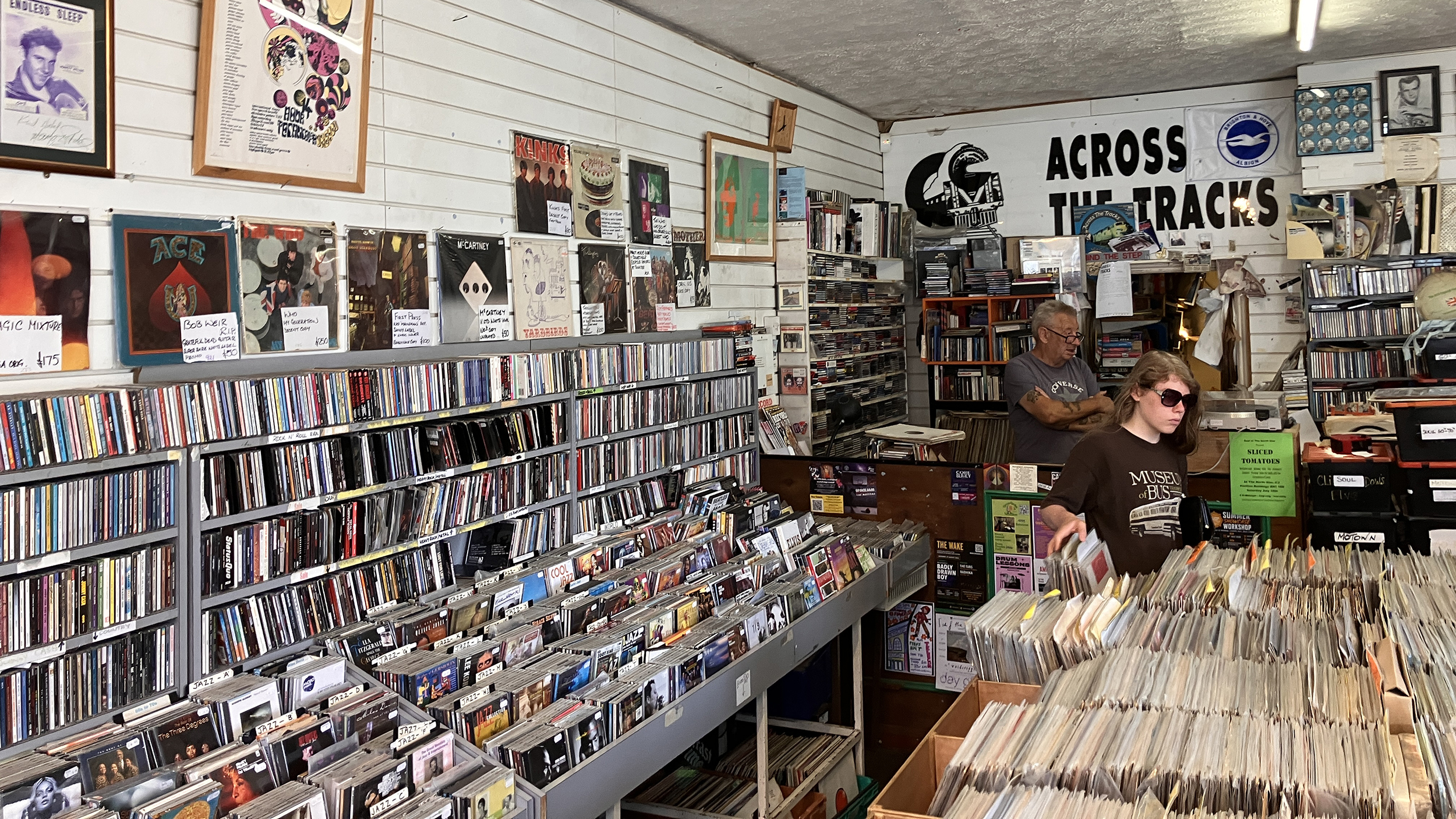 Innenraum eines dicht gefüllten Plattenladens mit Regalen voller CDs und Schallplatten; zwei Personen stehen am Verkaufstresen.