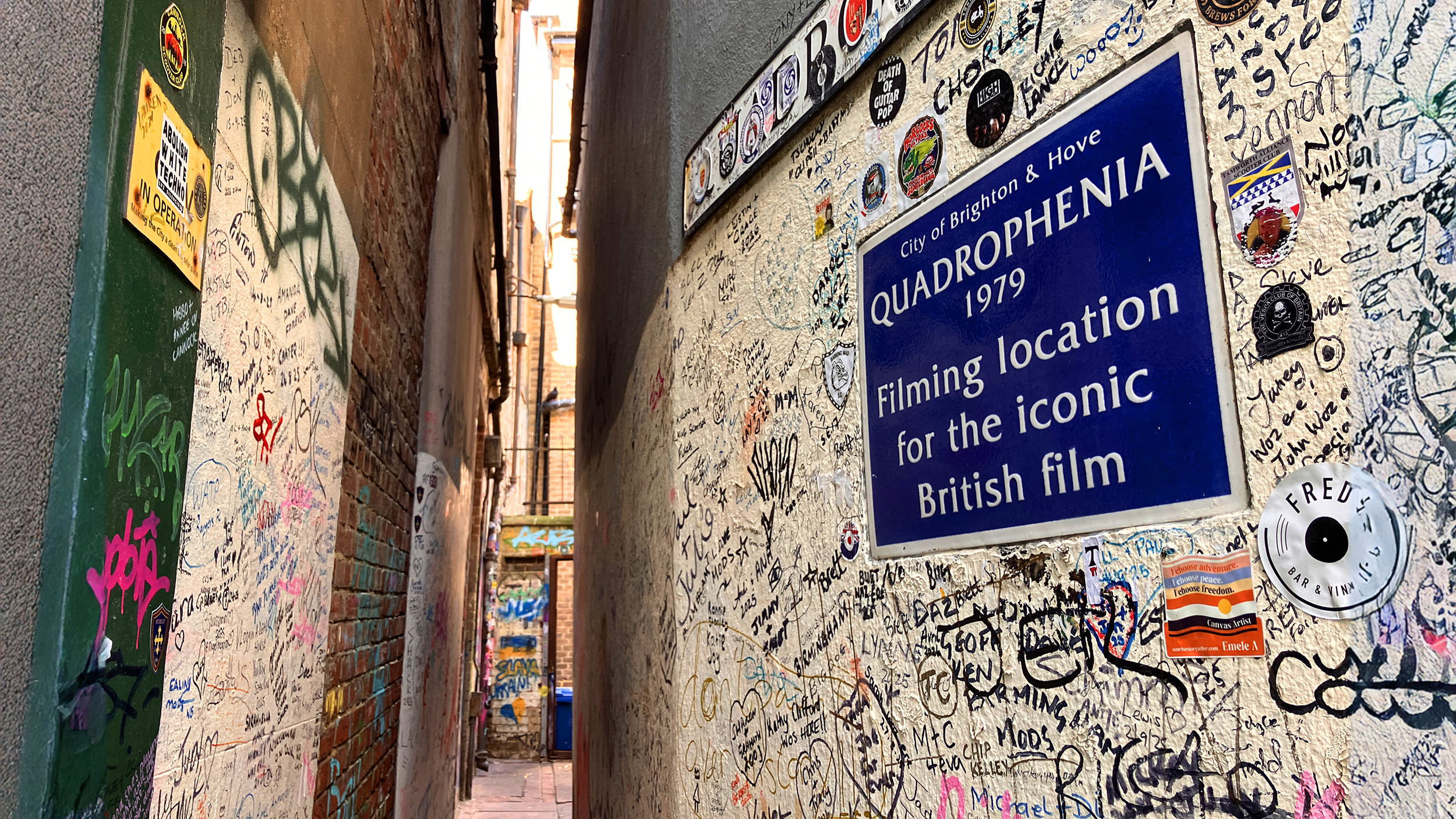 Enge, mit Graffiti, Aufklebern und handschriftlichen Botschaften bedeckte Gasse; an der rechten Wand hängt ein blaues Schild mit der Aufschrift „Quadrophenia 1979“.