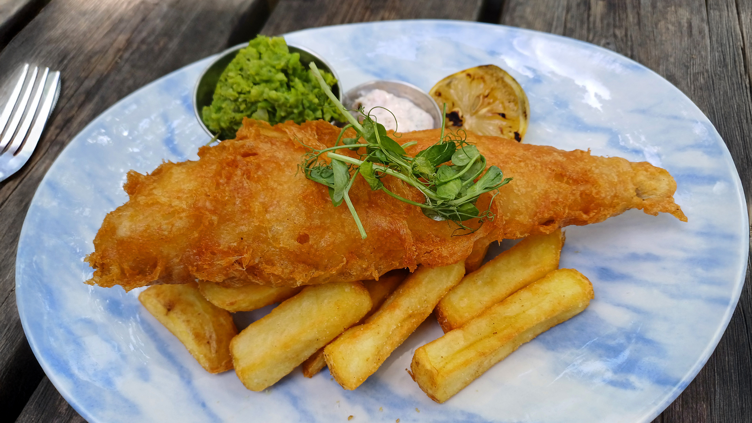 Teller mit paniertem, gebratenem Fisch auf dicken Pommes, dazu Erbsenpüree, Sauce, Zitrone und frische Kresse.