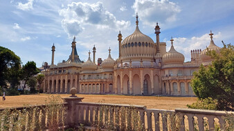 Royal Pavilion in Brighton mit zahlreichen Kuppeln und Türmchen hinter einer niedrigen Steinbalustrade und trockener Rasenfläche.