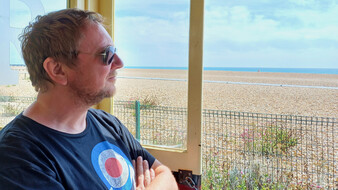 Blick aus einer cremefarbenen Strandbahn auf die Gleise, die Strandpromenade und den Kiesstrand am Meer.