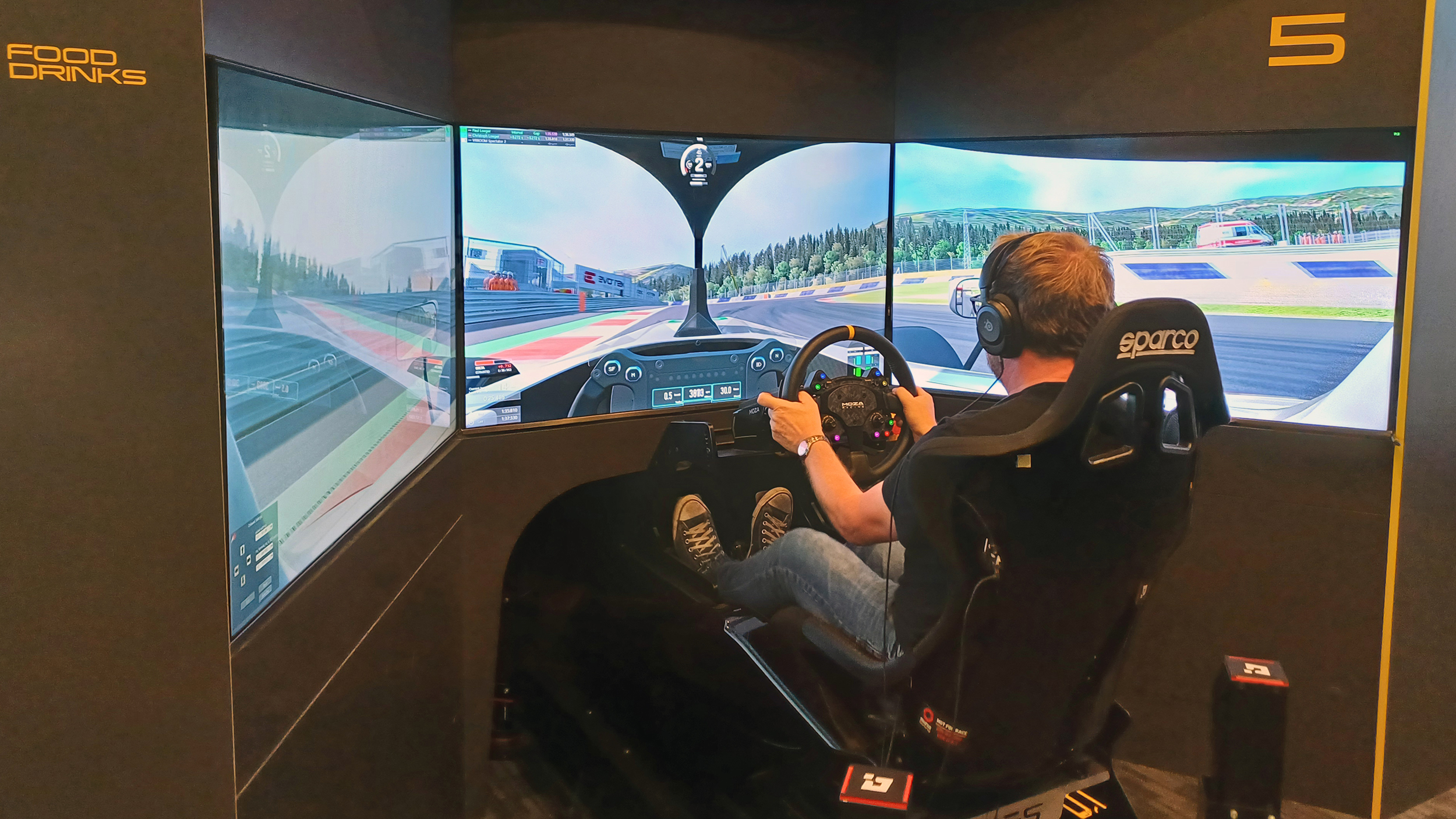 Mann sitzt in einem Rennsimulator mit drei Monitoren, Lenkrad und Pedalen; auf den Bildschirmen ist eine virtuelle Rennstrecke zu sehen.