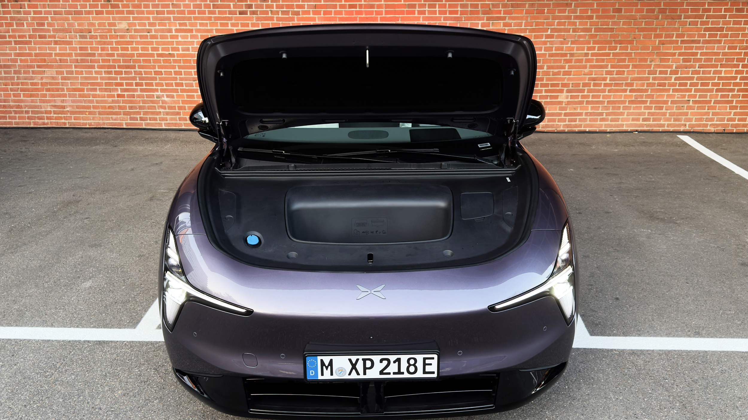 Frontansicht eines violetten SUV mit geöffneter Motorhaube und schwarzer Abdeckung mit mittigem Staufach vor einer Ziegelwand.
