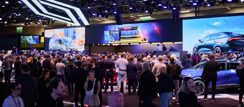 Viele Menschen auf einer Automesse vor einer Bühne mit Bildschirmen.