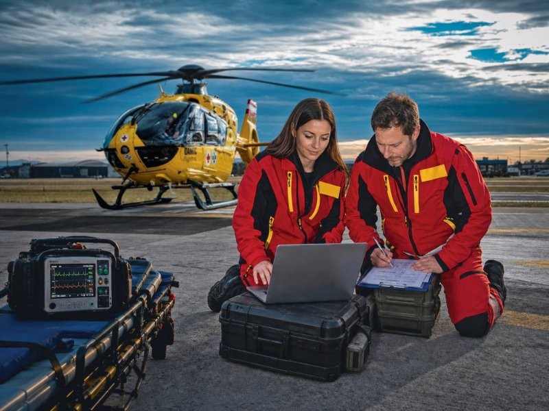 Eine Notärztin und ein Flugretter erfassen Daten auf einem Laptop. Dahinter ist ein Hubschrauber zu sehen.
