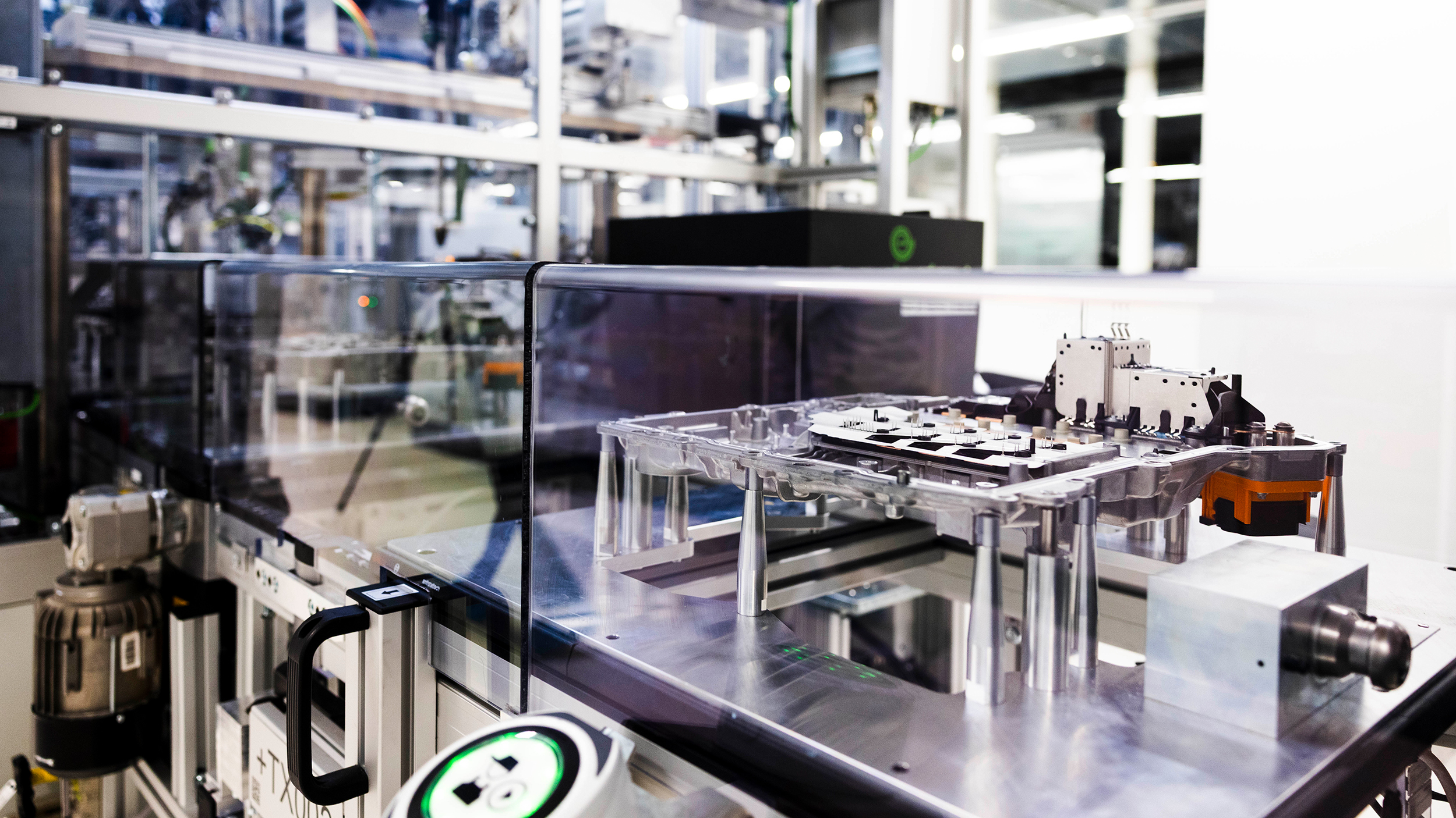 Offenes Metallgehäuse mit elektronischen Bauteilen und orangefarbenem Anschluss auf einer Halterung hinter einer transparenten Schutzabdeckung.