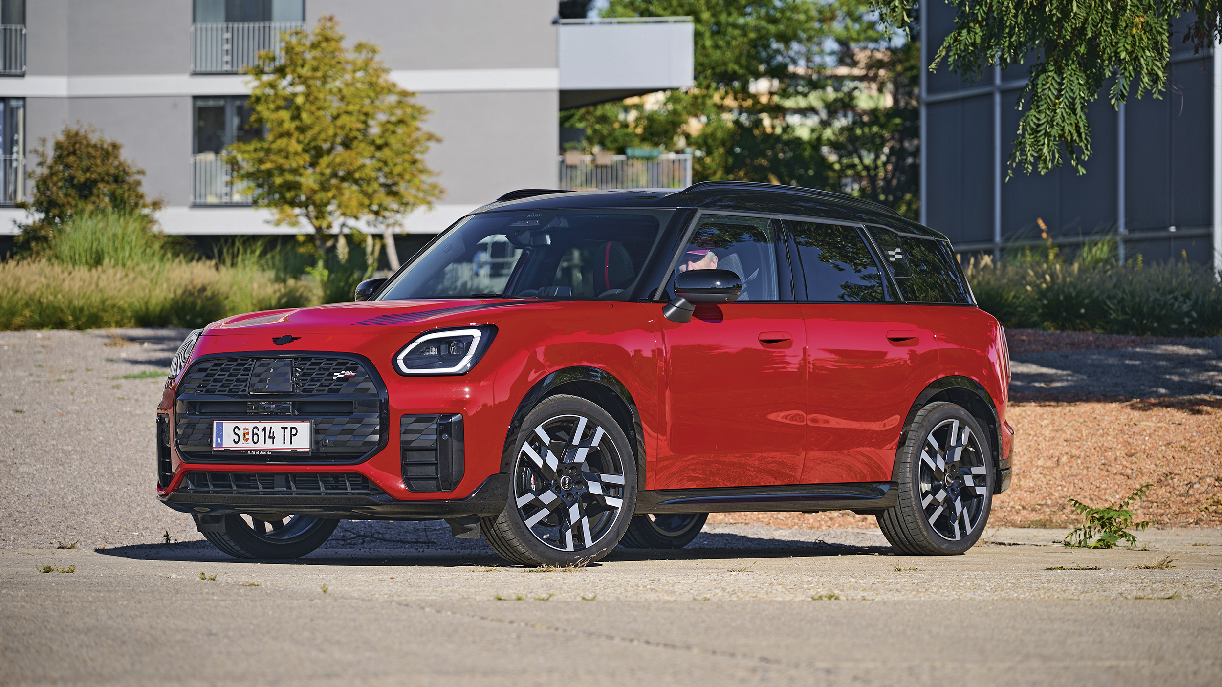 Roter MINI Countryman mit schwarzem Dach, schwarzen Radlaufverkleidungen und schwarz-silbernen Felgen schräg von vorn auf einem asphaltierten Platz.