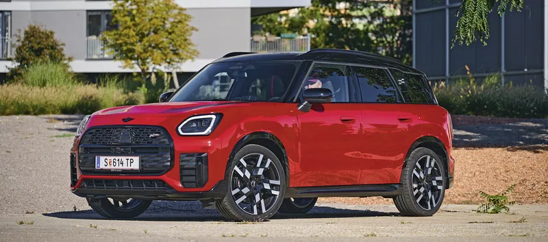 Roter MINI Countryman mit schwarzem Dach, schwarzen Radlaufverkleidungen und schwarz-silbernen Felgen schräg von vorn auf einem asphaltierten Platz.